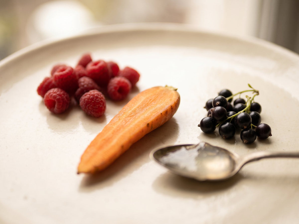 Raspberries, a halved carrot, blackcurrants and a spoon on a warm ivory surface