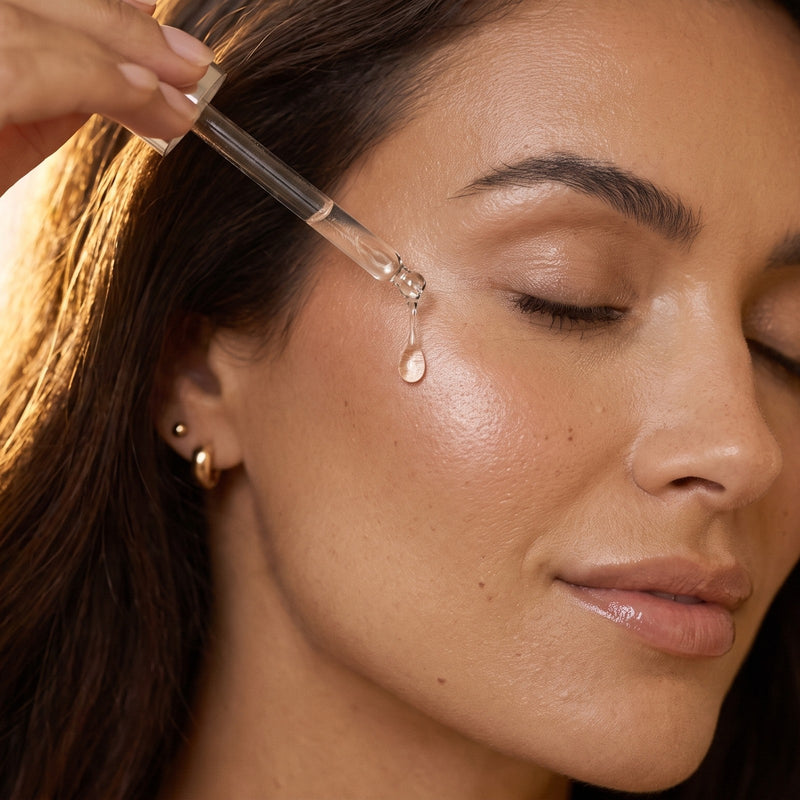 A drop of serum falling from a glass dropper onto a woman's cheekbone in morning light